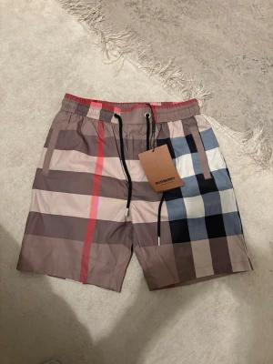 Burberry shorts - Snygga shorts från Burberry med klassiskt rutigt mönster i beige, svart, vit, blå och röd. Shortsen har elastisk midja med svart snörning och sidofickor. Perfekta för en trendig och avslappnad stil.