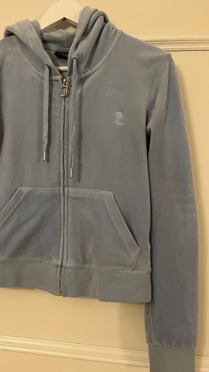 Ljusblå  hoodie Juicy Couture - Snygg ljusblå hoodie i mjuk velour från Juicy Couture. Dragkedja framtill, två stora fickor och klassisk huva med dragsko. Liten broderad logga på bröstet. Perfekt för en chill och trendig look.