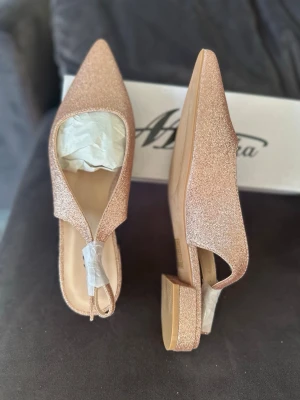 Glittriga roséguldiga pumps. Mulanka - Säljer ett par eleganta pumps / Slingbacks i roséguld med glittrig yta och spetsig tå från Mulanka. Materialet är syntet med glitterfinish. Klacken är på ca 2.5 cm och längden på skon är ca 27-27.5 cm. Älskar skorna men tyvärr trodde att de var i helt guld istället  för roséguld. Helt nya och såklart oanvända.