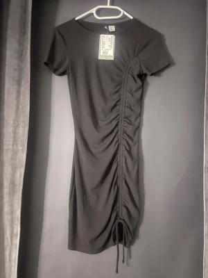 Svart Klänning - Säljer en svart klänning från H&M med snörning längs sidan som ger en snygg rynkad effekt. Klänningen har korta ärmar. Enkel och trendig modell. 