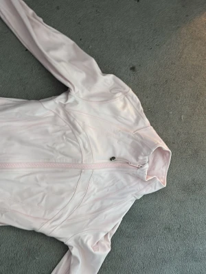 Ljusrosa träningsjacka från Lululemon - Säljer en ljusrosa träningsjacka från Lululemon med figurnära passform och hel dragkedja framtill. Jackan har långa ärmar och är tillverkad i ett stretchigt syntetmaterial som passar perfekt till träning eller uppvärmning.