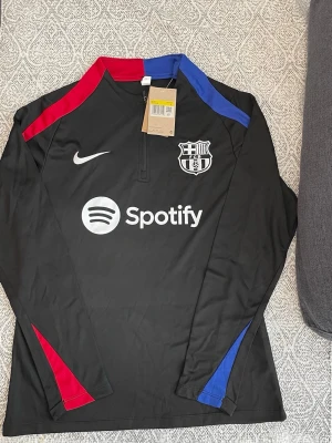 Svart Nike FC Barcelona träningströja - Svart långärmad träningströja från Nike med FC Barcelona-logga och Spotify-tryck på bröstet. Tröjan har dragkedja vid halsen och detaljer i rött och blått på axlar och ärmar. Perfekt för fotbollsträning eller som supporterplagg.