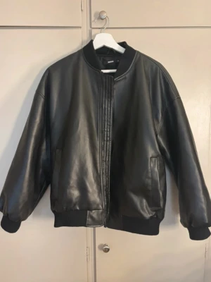 Svart skinnjacka i bombermodell - Säljer en svart skinnjacka från Bikbok i bombermodell. Jackan har ribbade muddar vid ärmslut och midja, två sidofickor och en stilren, minimalistisk design utan synliga knappar eller dragkedja framtill.