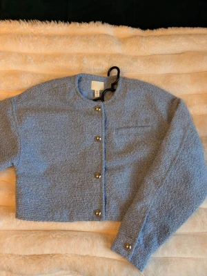 Ljusblå bouclé cardigan från H&M - Trendig ljusblå cardiganjacka från H&M i bouclétyg. Helt ny, oanvänd. Kort, boxig modell med rund halsringning och dekorativa silvriga knappar framtill samt på ärmsluten. Jackan har en bröstficka och är helfodrad för extra komfort.