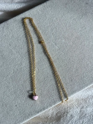 Guldhalsband med rosa hjärthänge - Sött halsband i guldfärgad metall med ett litet rosa hjärta som hänge. Kedjan är tunn och elegant, perfekt för att ge en gullig touch till din outfit. Hjärtat har en mjuk pastellrosa ton och sitter fast i en enkel guldfärgad kedja.