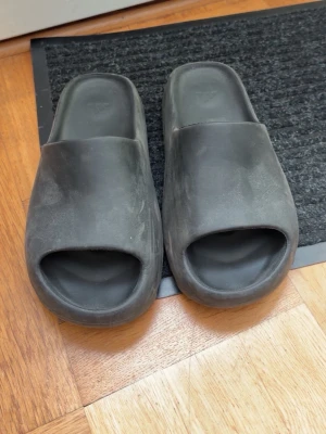 Svarta yeezy slides - Säljer ett par svarta Yeezy slides i mjukt gummi med öppen tå och bred rem över foten. Minimalistisk design med Adidas-logga på innersulan. Perfekta för chill dagar hemma eller till stranden. Jag har använt dom cirka två gånger och därför har dem blivit väldigt dammiga som man ser på tofflorna. 