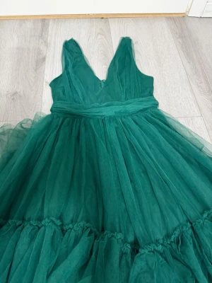 Grön långklänning med volanger - Säljer en fantastisk grön långklänning i tyll med djup v-ringning och breda axelband. Klänningen har flera lager volanger på kjolen som ger en riktigt maffig look. Perfekt för dig som vill sticka ut på bal eller fest! Materialet känns lätt och luftigt.