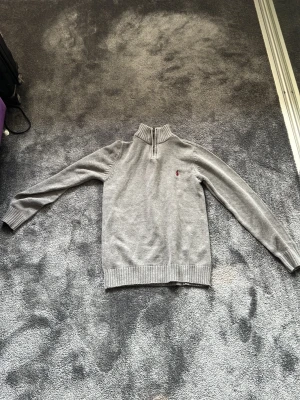 Grå stickad tröja med halv dragkedja - Snygg grå stickad tröja från Polo Ralph Lauren med halv dragkedja och hög krage. Tröjan har långa ärmar, ribbstickade muddar och en liten röd logga broderad på bröstet. Perfekt för lager på lager och enkel att matcha.