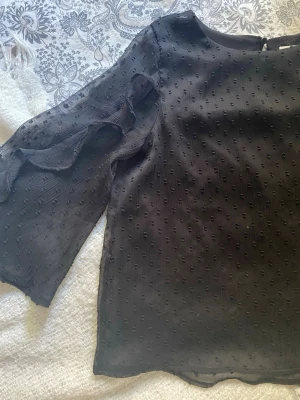 Svart blus med volangärm - Snygg svart blus i transparent material med diskret prickig struktur. Blusen har långa ärmar med volangdetaljer längs sidorna och rund halsringning. 