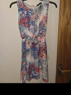 Blommig sommarklänning i blått och rött - Snygg kort blommig klänning med tropiskt mönster i blå, röd och vit. Ärmlös modell med rund halsringning och resår i midjan för en bekväm passform. Perfekt för varma dagar och somriga tillfällen.