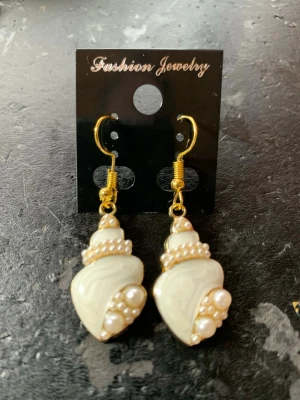 Snäckformade örhängen med pärlor - Unika örhängen i snäckform med guldfärgade krokar. Örhängena är dekorerade med små vita pärlor som ger en elegant och trendig look. Perfekta för dig som vill sticka ut med accessoarer inspirerade av havet.