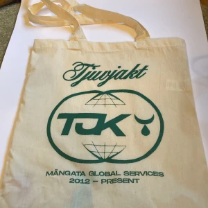 Tjuvjakt tygkasse med tryck - Beige tygkasse med grönt tryck från Tjuvjakt. Väskan har två långa axelband och motivet inkluderar texten 'Tjuvjakt', 'TJK' och 'Mångata Global Services 2012 - Present'. Perfekt för att bära böcker eller matvaror. Kommer även med 2 st 2 för 1 ingångar på Sunset festival i Uppsala om man vill ha, då ökas även priset.