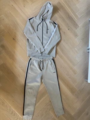 Beige tracksuit set från BOSS - Snygg beiget tracksuit set från BOSS med svarta sidoremsor och logotyp på benet och armarna. Perfekt för en avslappnad och trendig look.