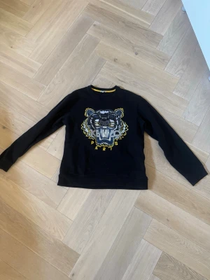 Svart Kenzo sweatshirt med tigertryck - Cool svart sweatshirt från Kenzo med stort tigertryck i grått, gult och vitt på bröstet. Perfekt för dig som vill sticka ut med ett ikoniskt motiv.