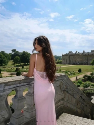 Ljusrosa långklänning med spetsdetalj - Elegant ljusrosa långklänning med tunna axelband och vacker spetsdetalj i ryggen. Klänningen har en figurnära passform och lätt rynkning vid midjan för en smickrande siluett. Perfekt för sommarens alla tillfällen.