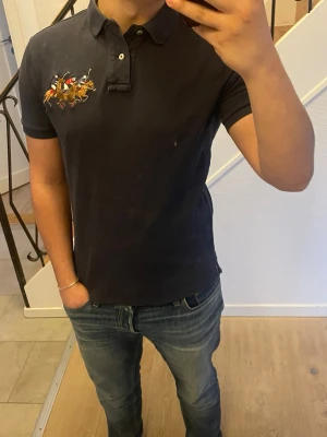 Mörkblå pikétröja från Polo Ralph Lauren - Mörkblå pikétröja från Polo Ralph Lauren med broderat polospel-motiv på bröstet och röd siffra på ärmen. Klassisk krage med knappar och korta ärmar. Perfekt till jeans för en avslappnad men snygg stil.