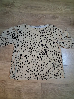 Beige blus med svart mönster - Trendig beige blus med svart abstrakt prickigt mönster och puffiga korta ärmar. Blusen har rund halsringning och rak passform. Perfekt för dig som vill ha en cool och modern look.