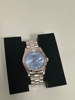 Silverfärgad klocka från Lotus - Stilren klocka från Lotus i silverfärg med länkarmband i metall. Urtavlan är ljusblå med datum- och veckodagsvisning samt markerade index med små stenar. Klassisk design som passar till många olika stilar. Säljer denna eftersom jag har köpt en annan klocka.