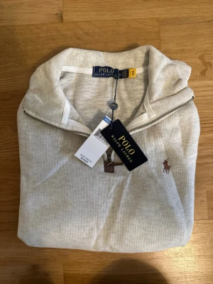 Ljusgrå tröja från Polo Ralph Lauren - Ljusgrå långärmad tröja från Polo Ralph Lauren med halv dragkedja och klassisk krage. Tröjan har det ikoniska broderade logotypen på bröstet och är tillverkad i mjukt material. Perfekt för en stilren och avslappnad look.