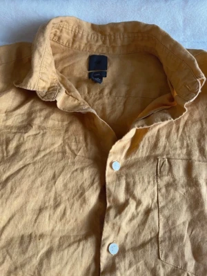 Gul kortärmad linneskjorta - Säljer en beige/gul (mer gul) kortärmad skjorta från H&M i linne. Skjortan har klassisk krage, vita knappar framtill och en bröstficka. Perfekt för varma dagar och enkel att styla till olika outfits. Har bundle rabatter, den är helt ny. Storlek M. 