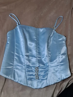 Ljusblå satin korsett med strassdetaljer - Superfin ljusblå korsettopp i satin med smala axelband och glittriga strassdetaljer framtill. Toppen har figurnära passform och dragkedja i ryggen. Perfekt för dig som vill sticka ut med en trendig look. Storlek 36/38 mer åt 38