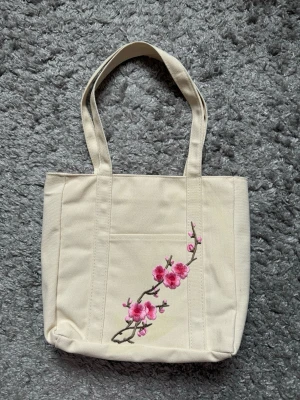 Beige tygkasse med broderade blommor - Snygg beige shoppingväska i tyg med broderade rosa körsbärsblommor på framsidan. Väskan har två långa handtag och en fyrkantig form, perfekt för att bära med sig allt man behöver. Materialet är slitstarkt tyg och designen är både söt och stilren.