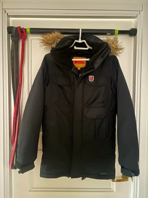 Svart parkas från Fjällräven - Snygg svart parkas från Fjällräven med avtagbar huva och fuskpäls. Jackan har flera praktiska fickor framtill och klassisk Fjällräven-logga på bröstet. Perfekt för kalla dagar och riktigt skön att bära. Materialet är slitstarkt och vindtätt.