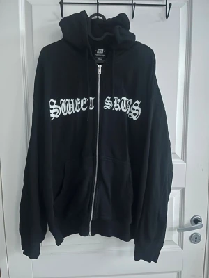 Svart hoodie från Sweet Skys - Cool svart hoodie från Sweet Skys med vit text på bröstet i gotisk stil. Tröjan har dragkedja, stor huva och två rymliga fickor framtill. Materialet är mjukt och bekvämt, perfekt för att chilla eller hänga med kompisar.