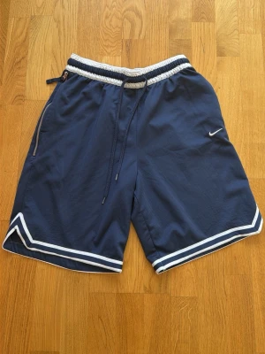 Marinblå Nike shorts med snörning - Snygga marinblå shorts från Nike med vita detaljer och snörning i midjan. Shortsen har dragkedja på ena fickan och elastisk midja för skön passform. Perfekta för träning eller chill dagar. Klassisk Nike-logga på benet och sportig look.