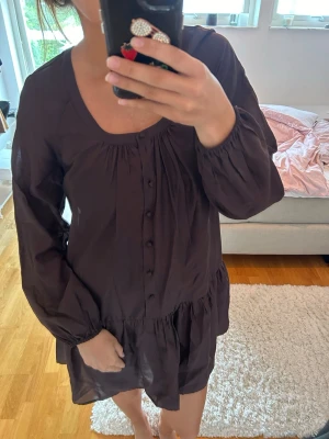 Mörk brun kortklänning med knappar - Säljer en mörkbrun kort klänning med långa ärmar och knappar framtill. Klänningen har en lös passform, rundad halsringning och volang nedtill. Perfekt för dig som gillar en enkel men ändå snygg stil.