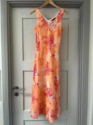 Blommig långklänning i orange och rosa - Säljer en somrig långklänning med blommigt mönster i orange, rosa och vitt. Klänningen har v-ringning, ärmlös design och lätt utsvängd nederdel. Perfekt för varma dagar och har en mjuk, följsam passform.