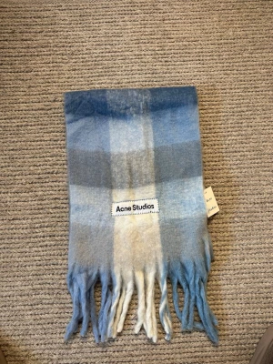Rutig halsduk från Acne Studios - Supersnygg rutig halsduk från Acne Studios i mjuk ullblandning. Färgskalan går i blått, grått och beige med stora rutor och långa fransar i ändarna. Perfekt accessoar för att lyfta din outfit med en trendig touch.