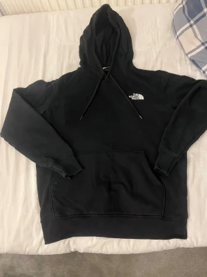 Svart hoodie från The North Face - Säljer en svart hoodie från The North Face med klassisk logga på bröstet. Hoodien har huva med dragsko, stor magficka och ribbade muddar vid ärmar och nederkant. Perfekt för en chill och avslappnad stil. Materialet är mjukt och bekvämt, troligen bomullsblandning.
