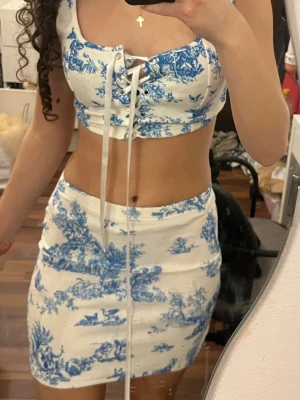 Vitt och blått set med snörning - Säljer ett snyggt tvådelat set med crop top och kjol i vitt med blått toile-mönster. Toppen har snörning framtill och breda axelband, kjolen är figurnära och kort. Perfekt för dig som gillar trendiga och feminina plagg.