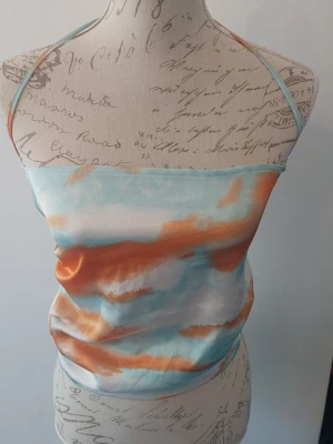 Färgglad satinlinnetopp med knyt - Trendig linnetopp i satin med ljusblå, orange och vita toner. Toppen har tunna axelband som knyts i nacken och öppen rygg för en cool och somrig vibe. 