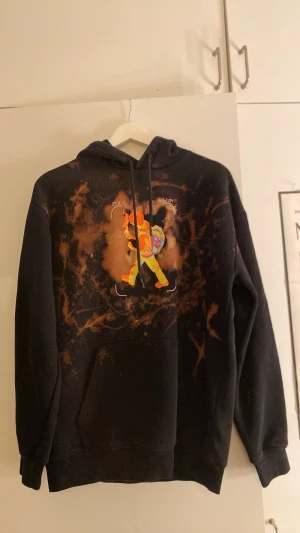 Svart hoodie med färgglatt tryck - Cool svart hoodie med unikt färgstänkt mönster och ett stort färgglatt tryck av en person med ryggsäck på bröstet. Hoodien har huva med dragsko och en stor magficka. Perfekt för dig som vill sticka ut med streetstyle vibes.