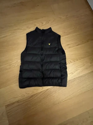 Svart dunväst från Lyle & Scott - Stilren svart dunväst från Lyle & Scott med gul logga på bröstet. Västen har hög krage, dragkedja framtill och breda horisontella kanaler. Perfekt för vardagen. Storlek 14-15 år.