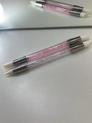 Silikonverktyg för nageldekoration - Två dubbelsidiga silikonverktyg för nageldekoration med transparent skaft fyllt av rosa och vita kristaller. Spetsarna är vita och tillverkade i mjuk silikon, perfekta för att forma gelé eller placera dekorationer på naglarna. Verktygen har silverfärgade metalldetaljer.