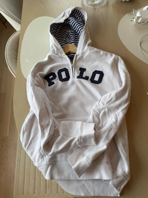 Vit hoodie från Polo Ralph Lauren - Vit hoodie från Polo Ralph Lauren med mörkblå POLO-text på bröstet. Huvan har ett snyggt marinblått och vitt randigt foder och dragkedja vid halsen. Klassisk känguruficka framtill och ribbade muddar vid ärmar och nederkant. Perfekt för en avslappnad och stilren look. Passar för storlek S.