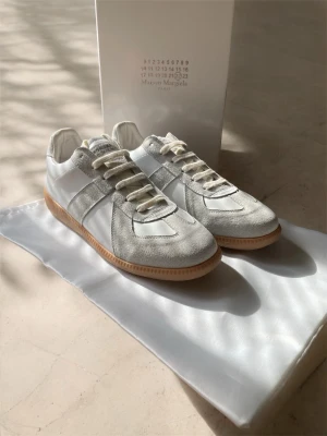 Maison Margiela vita och grå sneakers - Stilrena sneakers från Maison Margiela i vitt och ljusgrått med ovandel i läder och mocka. Klassisk låg modell med beige gummisula och vita snören. Perfekta för dig som gillar minimalistisk och clean stil.