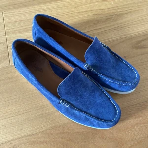 Blå mocka loafers från Polo Ralph Lauren - Snygga blå loafers i mocka från Polo Ralph Lauren med vita kontrastsömmar och vit sula. Klassisk design med rundad tå och diskreta detaljer vid hälen. Knappt använda, storlek 43