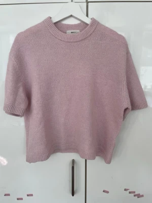 Ljusrosa stickad topp - Kortärmad stickad tröja i en mjuk ljusrosa nyans. Tröjan har rund halsringning och ribbade kanter vid ärmslut, hals och nederkant. Perfekt för dig som gillar en enkel men trendig look, jätte fin att stylea med ett vitt linne under och ett par ljusblå jeans!