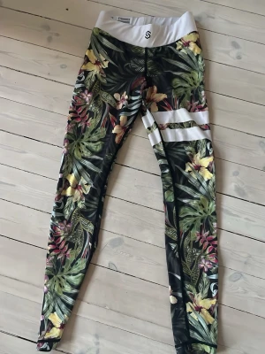 Stronger - Snygga leggings från Stronger med tropiskt blommönster. De har vita detaljer i midjan och två vita ränder på ena låret. Stretchigt material, perfekta för träning.
