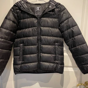 Svart pufferjacka från Champion - Snygg svart pufferjacka från Champion med dragkedja framtill och huva. Jackan har ett diskret Champion-tryck på bröstet och logotypdetaljer på huvan. Tillverkad i glansigt syntetmaterial och har två sidofickor. Perfekt för kalla dagar.