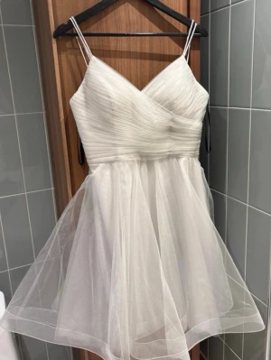 Vit kortklänning med snörning - Superfin vit kortklänning med tunna axelband och omlott-v-ringning. Klänningen har ett transparent, flowy tyllmaterial över kjolen och snörning i ryggen som ger en snygg detalj. Perfekt för dig som vill ha en elegant och modern look. Jag hade klänning på min konfirmationsfest.🥰