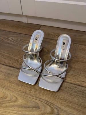 Silvriga strappy sandaler från Gina Tricot - Säljer ett par silvriga sandaler från Gina Tricot med fyrkantig tå och flera tunna remmar över foten. Klackhöjden är 9 cm. Endast använda en gång.