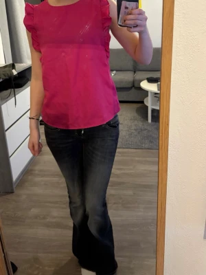 Rosa volangtopp med ärmlös design - Snygg rosa topp med volangdetaljer längs axlarna och ärmlös design. Toppen har rund halsringning och är tillverkad i ett lätt, slätt material som ger en fräsch look. Perfekt för dig som gillar färg och vill sticka ut lite extra.