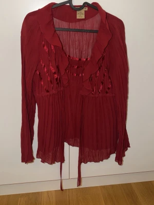 Vinröd plisserad blus från Notations - Snygg vinröd blus från Notations i plisserat tyg med volangkant och dekorativa satindetaljer framtill. Blusen är tillverkad i 100% polyester och har långa ärmar samt knytband nertill. Perfekt för dig som vill ha en romantisk och trendig look.