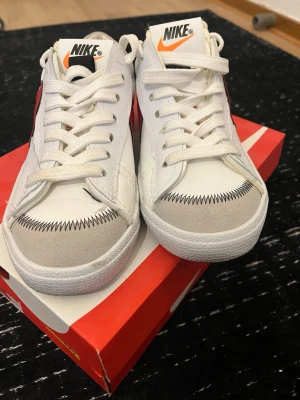 Nike SB Blazer Low vita/röda sneakers - Nike SB Blazer Low sneakers i vitt läder med röda detaljer och röd swoosh på sidan. Skorna har vit sula, vita skosnören och beige mockadetaljer vid tån. Klassisk Nike-logga på plösen och hälen. Perfekta för en clean och sportig stil.