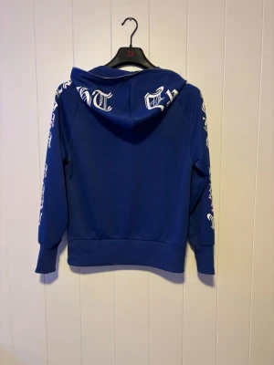 Blå hoodie med tryck på ärmar - Snygg blå hoodie med dragkedja och huva. Tryck i vit gotisk stil längs båda ärmarna och på huvan. Två fickor framtill och ribbade muddar vid ärmslut och nederkant. Perfekt för en streetwear-look och riktigt bekväm att bära. Pris kan diskuteras 